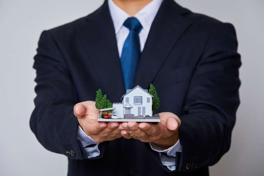 Un agent immobilier en costume tenant une maquette de maison avec jardin et voiture dans ses mains ouvertes, représentant une offre d'investissement immobilier.